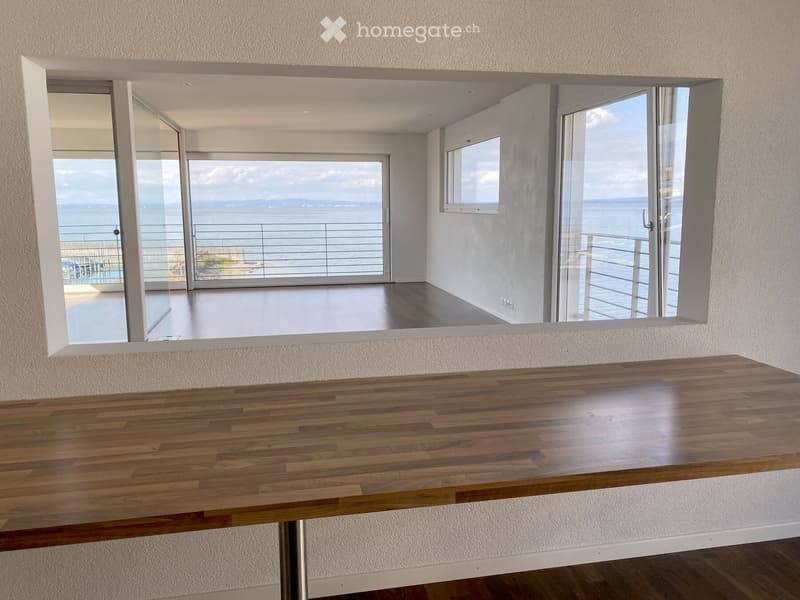A modern, minimalist room with a large wooden table, large windows offering a scenic ocean view, and a balcony accessible through sliding glass doors.