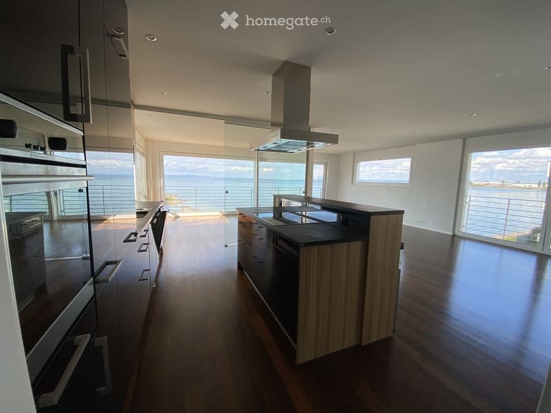 Modern kitchen with wooden flooring, stainless steel appliances, island, and expansive ocean view through large windows.