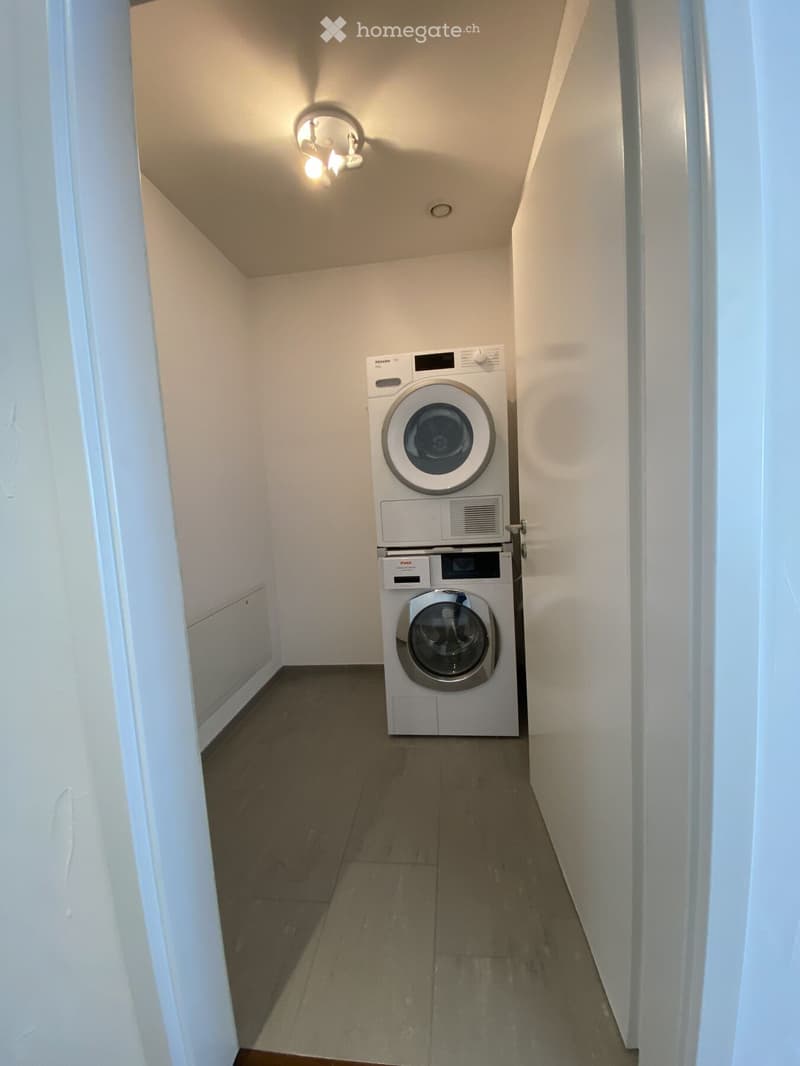 A narrow hallway with a stacked washing machine and dryer unit against the wall, white walls, and a ceiling light fixture.