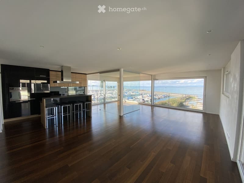 Modern kitchen with dark wooden flooring, sleek black cabinets, stainless steel appliances, and a large island with four stools. Large windows offer a scenic view of a marina and the sea.