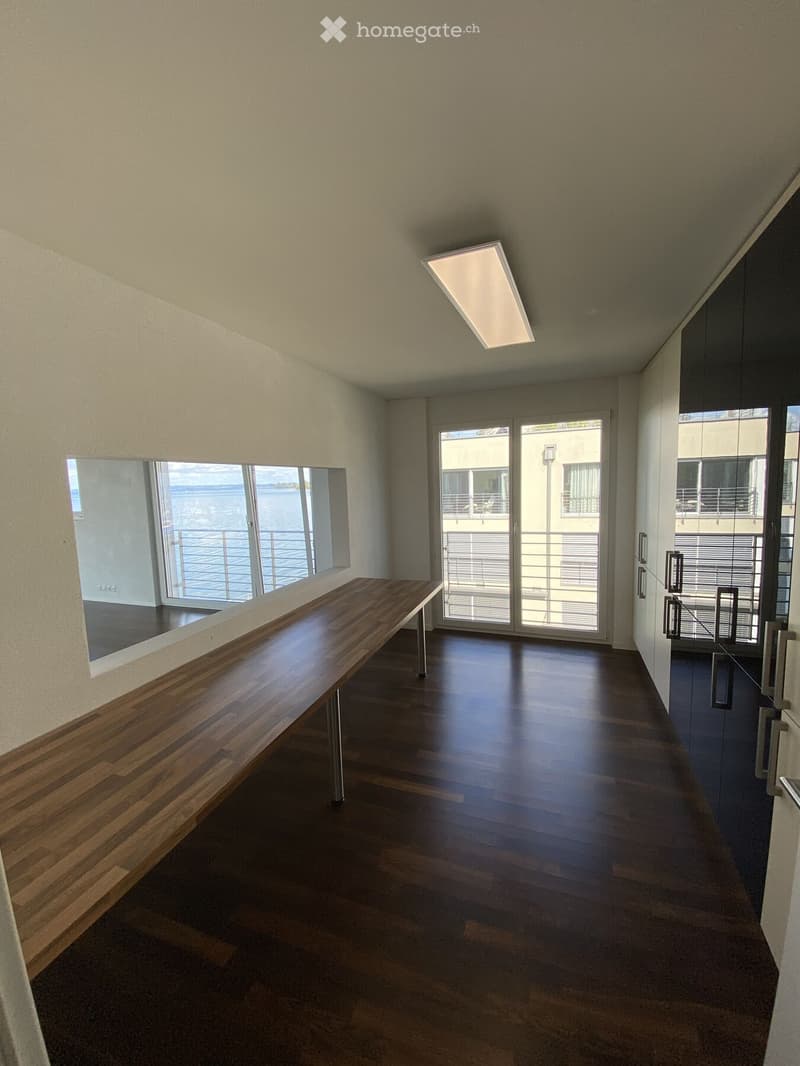 Modern hallway with wooden flooring, large windows, and a long wooden bench against the wall. The hallway features a sliding glass door leading to a balcony with a view of the ocean.