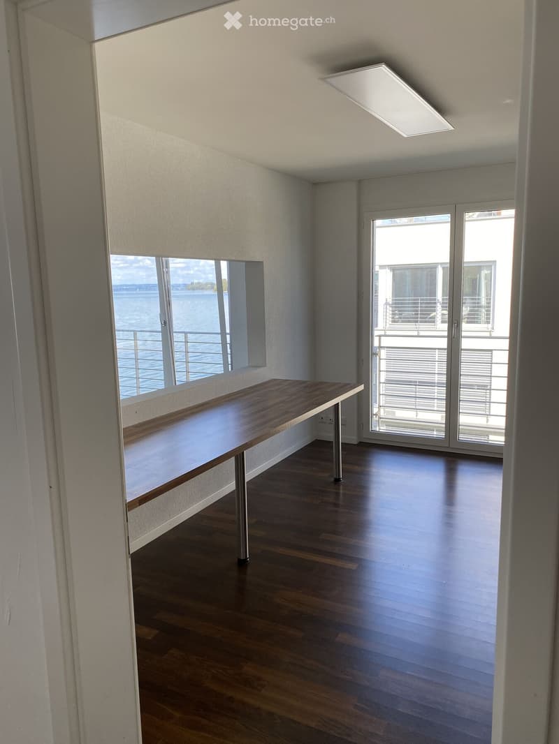 Bright, minimalist room with wooden flooring, a long wooden table, large windows offering a scenic view, and a sliding glass door leading to a balcony.
