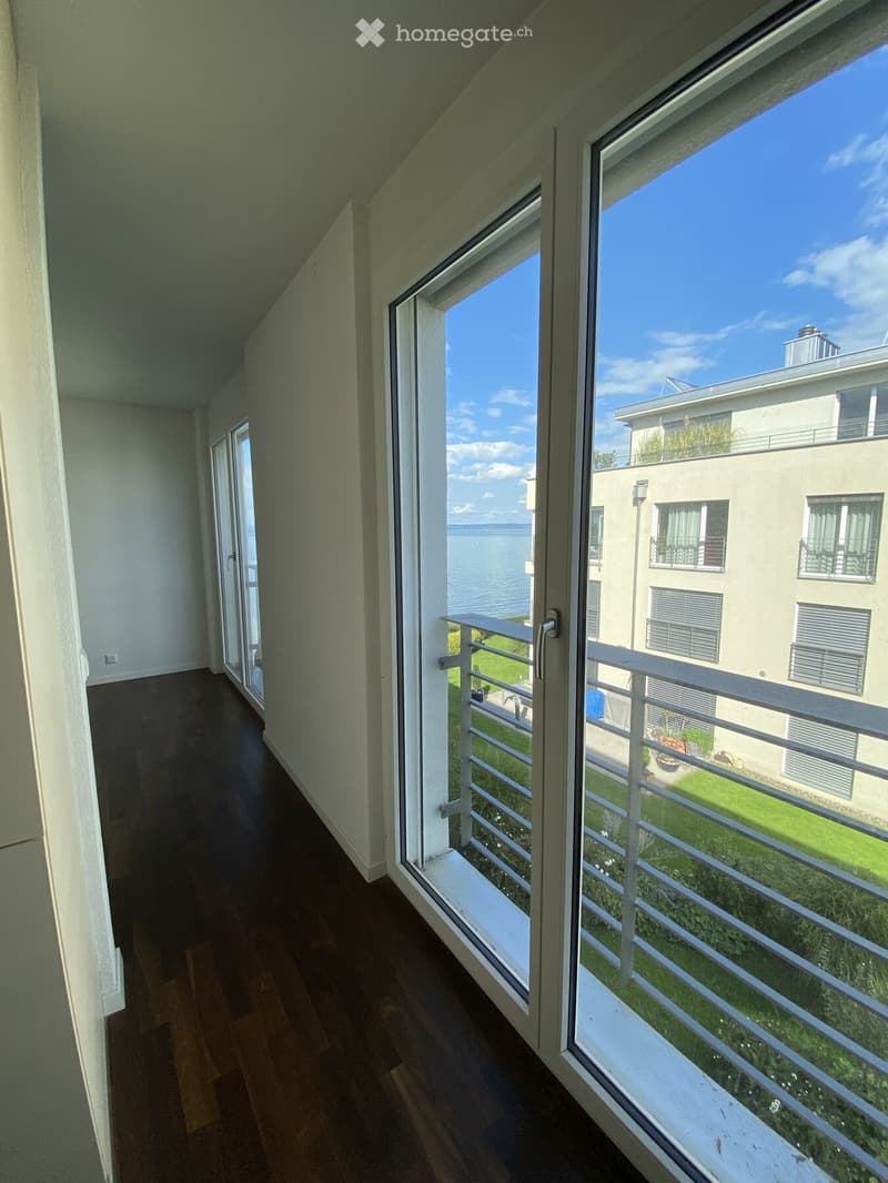 Balcony with wooden flooring, white railing, and large windows offering a view of the water and surrounding buildings.