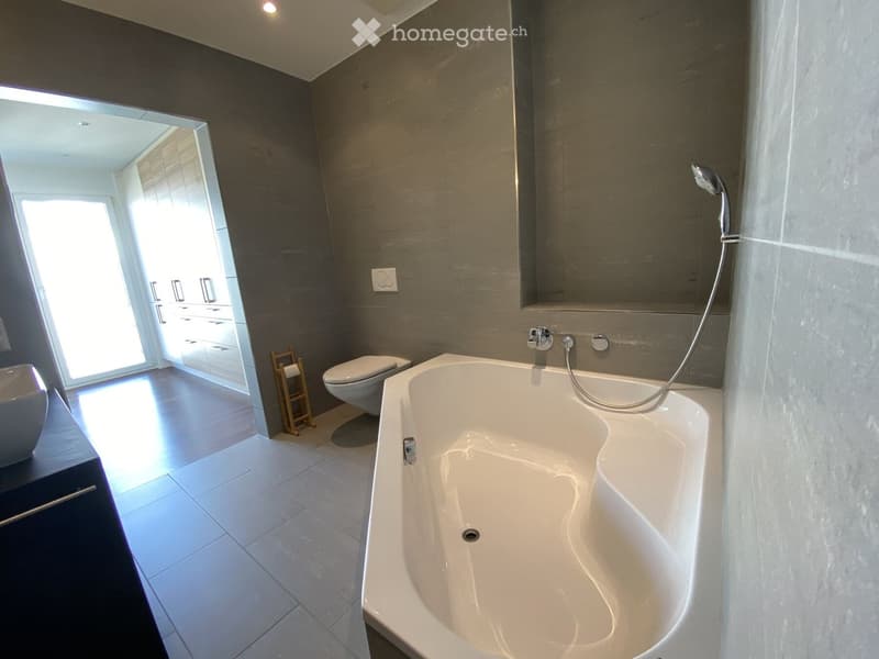 Modern bathroom with a large bathtub, toilet, sink, and a large mirror. The room features dark grey tiles and a wooden towel rack.