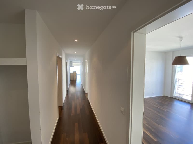 Empty hallway with wooden flooring, white walls, and recessed lighting. A pendant light is visible in the adjacent room.