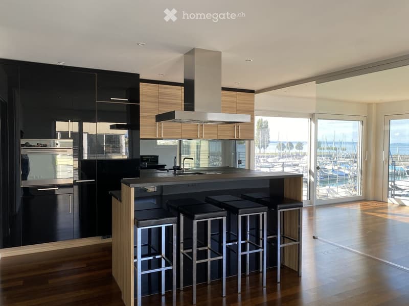 Modern kitchen with a central island, black and wood cabinets, stainless steel appliances, and a view of a marina through large windows.