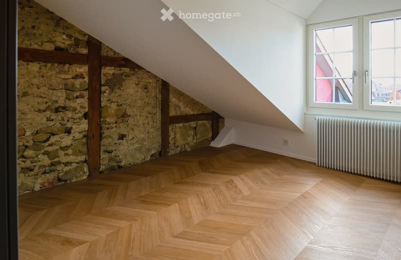 Empty attic room with wooden herringbone flooring, exposed stone walls, and large windows with white frames. A radiator is visible under the windows.