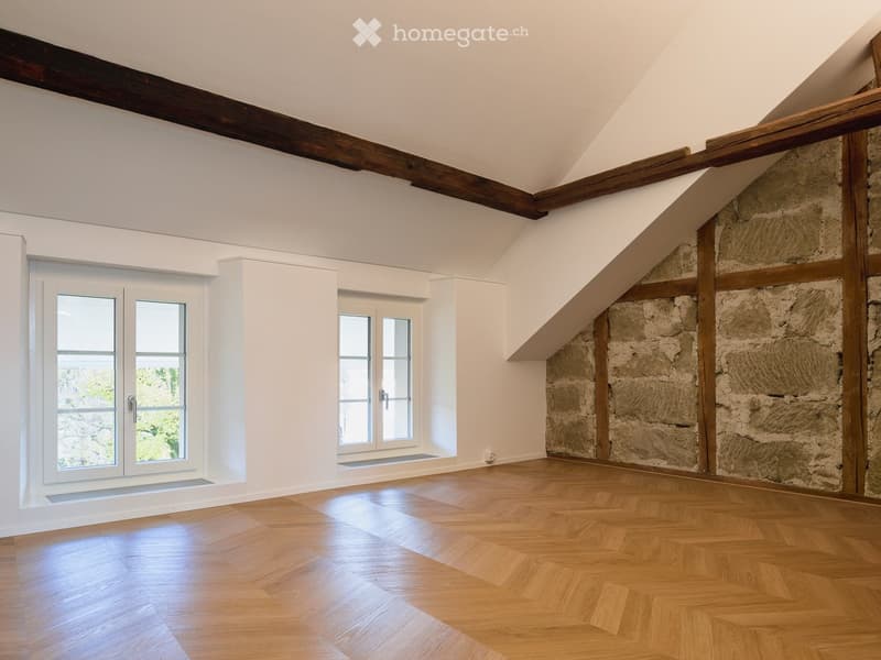 Bright, unfurnished room with wooden flooring, white walls, exposed wooden beams, and large windows offering natural light.