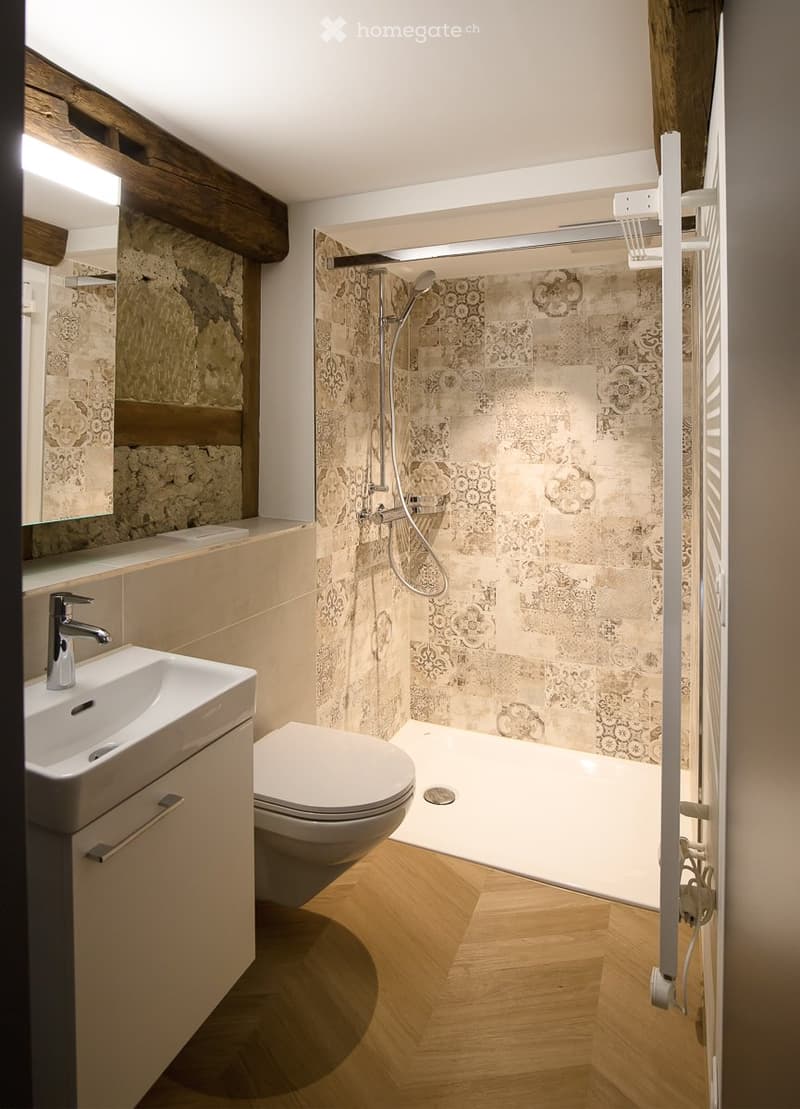 Modern bathroom with wooden flooring, a white sink with a chrome faucet, a toilet, a shower with a glass enclosure, and a decorative mirror above the sink.