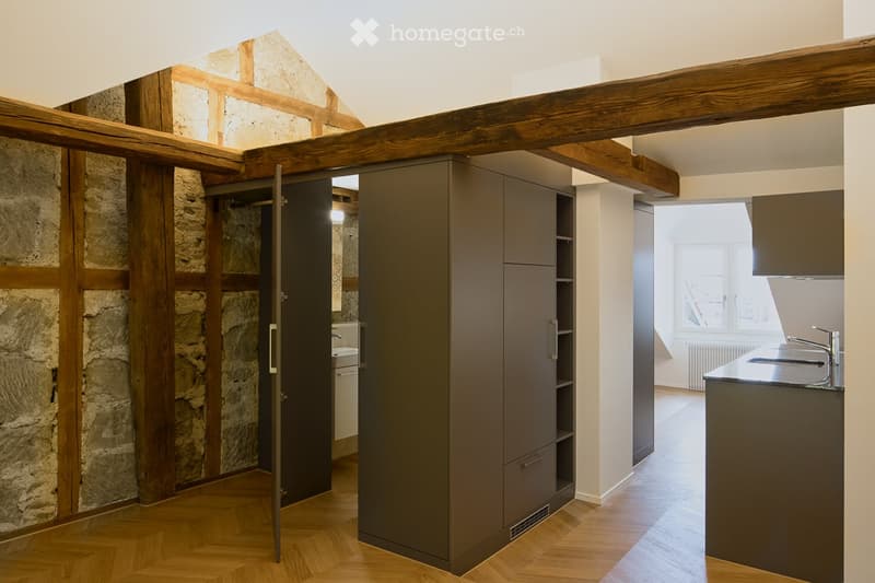 Modern kitchen with wooden herringbone flooring, exposed wooden beams, stone wall, grey cabinets, and a sink with a faucet.