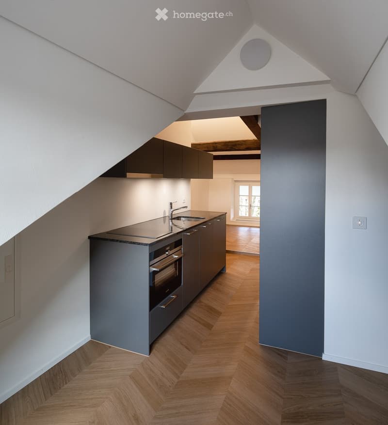 Modern kitchen with grey cabinets, stainless steel sink and oven, wooden herringbone flooring, and exposed wooden beams in the ceiling.