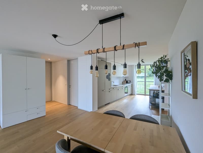 Open-plan living and dining area with a wooden dining table, chairs, white kitchen cabinets, pendant lights, and a plant in the corner.