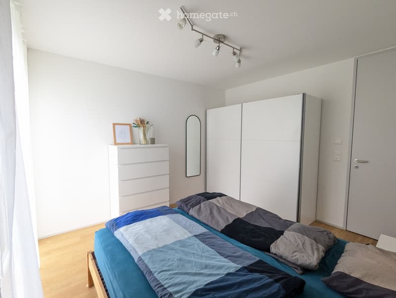 Modern bedroom with a double bed, blue and grey bedding, white wardrobe, mirror, nightstand with a picture frame and vase, and track lighting.