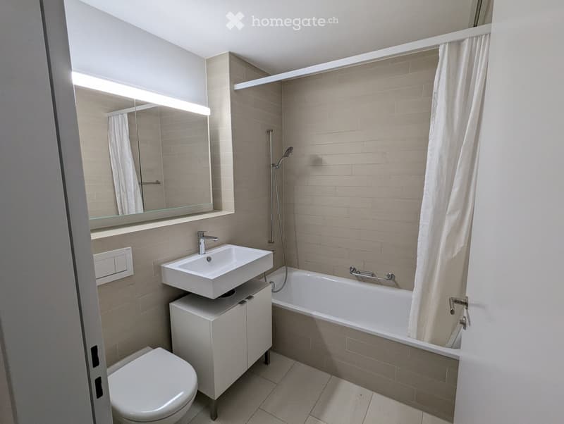 Modern bathroom with a white sink, toilet, bathtub, and large mirror. Beige tiles and a shower curtain are visible.