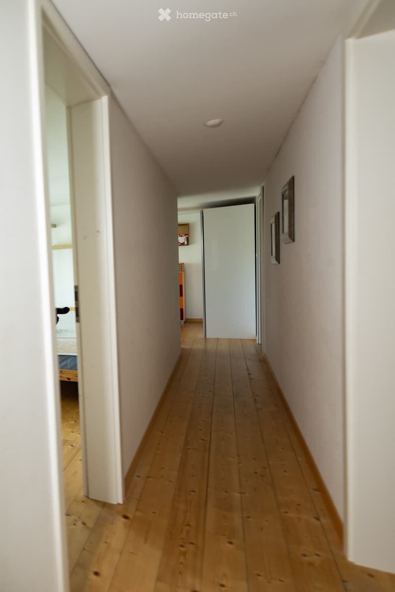 A narrow hallway with wooden flooring, white walls, and recessed lighting. Doors lead to adjacent rooms, and framed artwork is visible on the right wall.