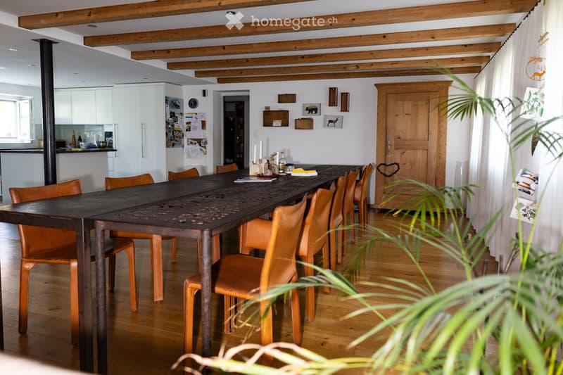 A spacious dining area with a long wooden table surrounded by orange chairs, exposed wooden beams on the ceiling, and a modern kitchen in the background.