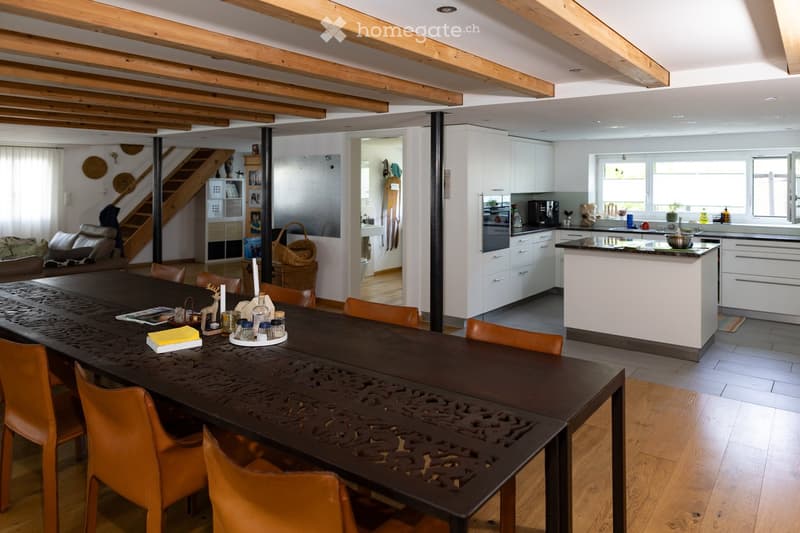 Open-plan living and dining area with exposed wooden beams, a long dark wooden dining table with orange chairs, a modern kitchen with white cabinets and a central island, and a comfortable living area with a sofa and decorative items.