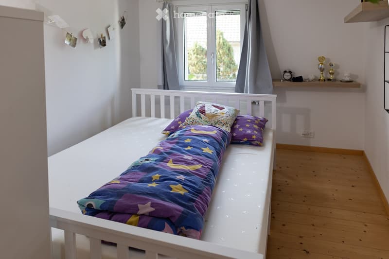 Cozy children's bedroom with a single bed, colorful star-patterned blanket, pillows, and a small shelf holding decorative items and a clock.