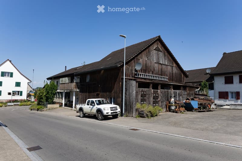 A large wooden barn with a pitched roof and satellite dish, situated on a street with neighboring houses and a parked truck.