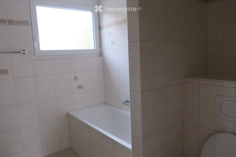Bathroom with a white bathtub, tiled walls, and a toilet. Features a window with a white frame and a towel rack on the wall.