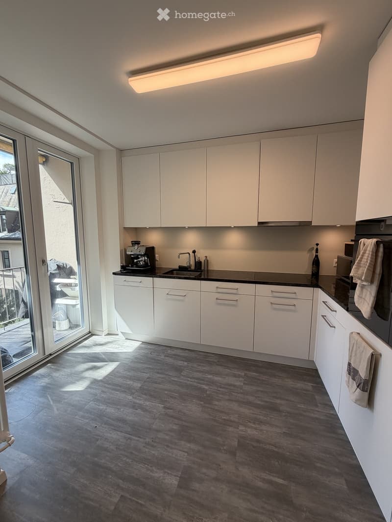 Modern kitchen with white cabinets, dark countertops, and wooden flooring. Features a coffee maker, sink, and oven.