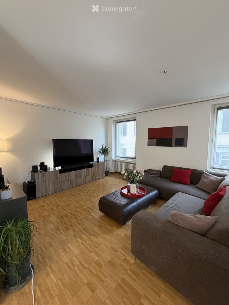 Modern living room with wooden flooring, large sectional sofa, coffee table, flat-screen TV on a sideboard, and decorative plants.