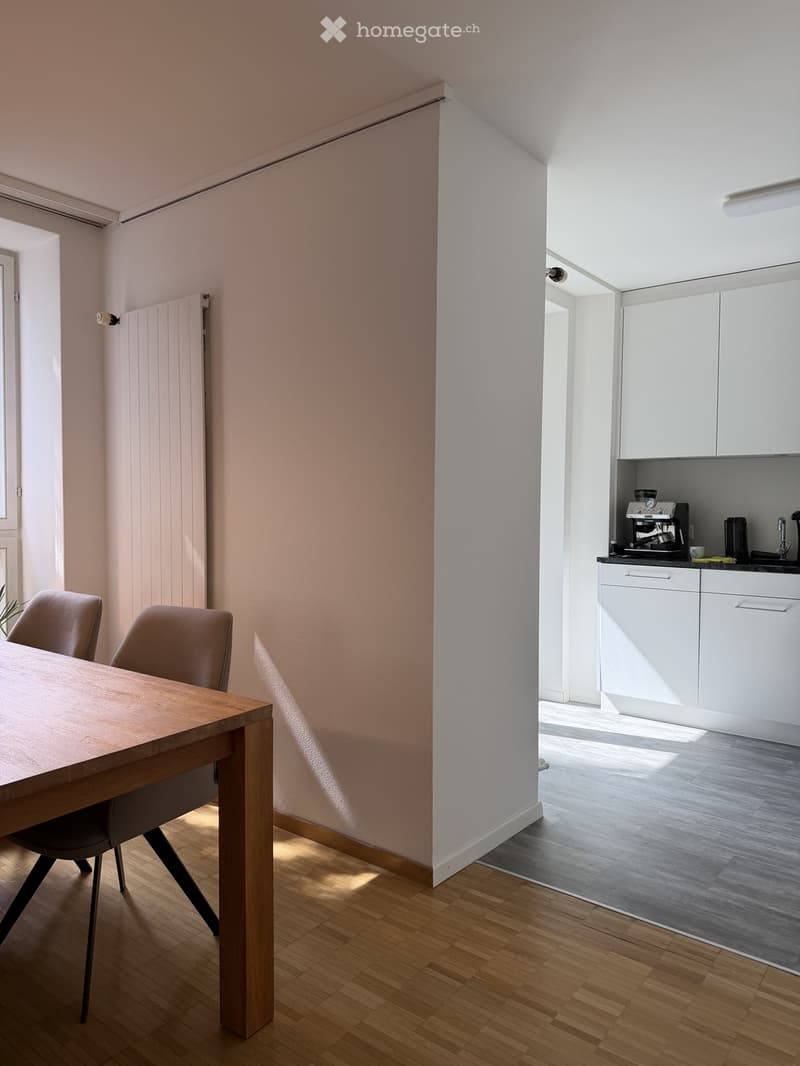 A modern, open-plan dining and kitchen area with light wooden flooring, a wooden dining table with two chairs, and a white kitchen with cabinets and a coffee machine.
