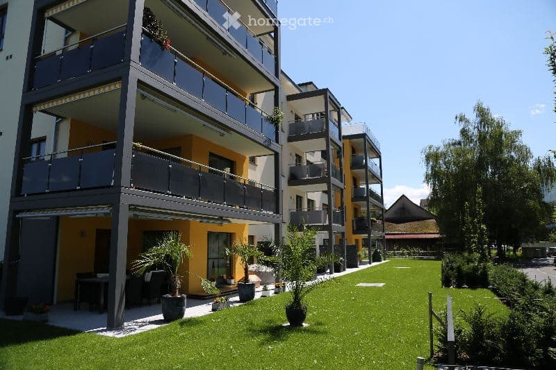 Modern apartment complex with multiple balconies, green lawn, potted plants, and trees.