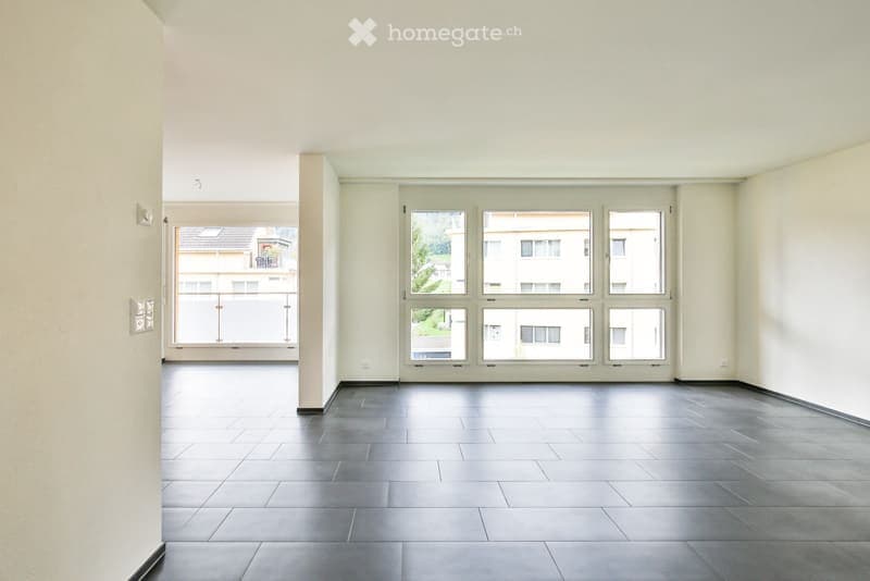 Empty room with large windows and a balcony door, featuring a dark tiled floor and white walls.