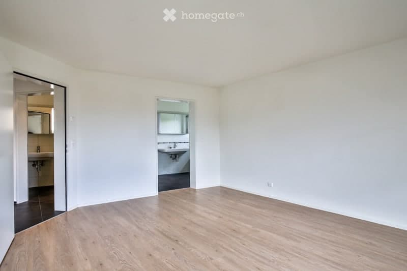 Empty room with light-colored wooden flooring and white walls. Two adjacent bathrooms are visible through open doorways, each equipped with a sink and mirror.