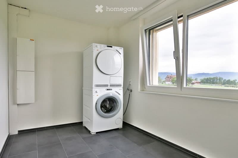 Empty room with a washing machine and dryer, white walls, dark tiled floor, and large windows with a scenic view.