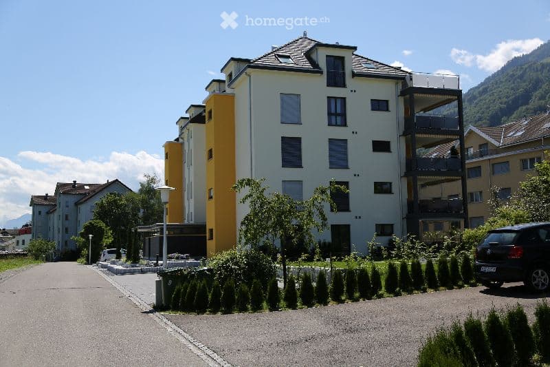 Modern multi-story residential building with white and yellow facades, multiple windows, balconies, and surrounding greenery.