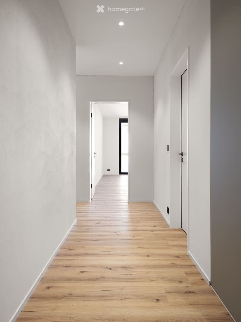 A modern, minimalist hallway with light wooden flooring, white walls, and recessed ceiling lighting. The hallway features several doors and a visible electrical outlet on the right wall.