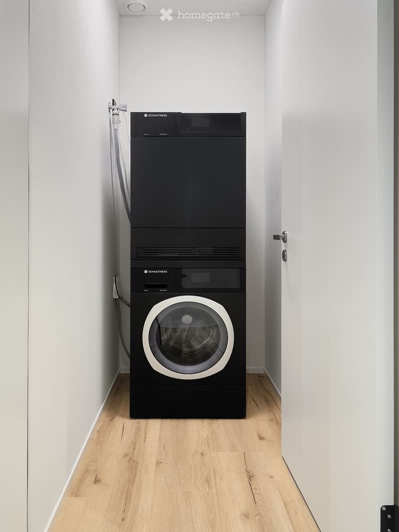 Modern, compact laundry room with light wooden flooring and white walls, featuring a stacked washer and dryer unit.