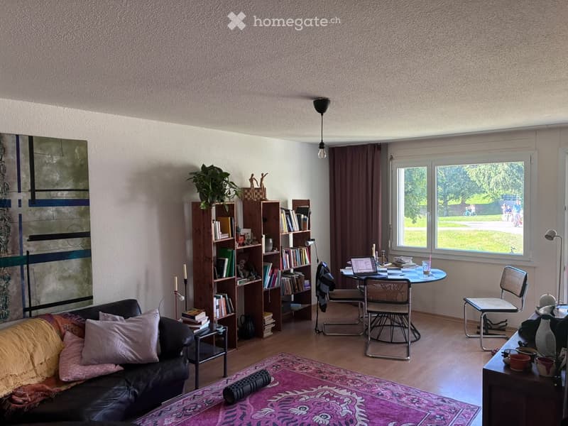 A cozy living room with a black leather sofa, a bookshelf filled with books, a round table with chairs, a laptop, and a plant on the windowsill. The room features large windows with curtains and a colorful rug on the floor.