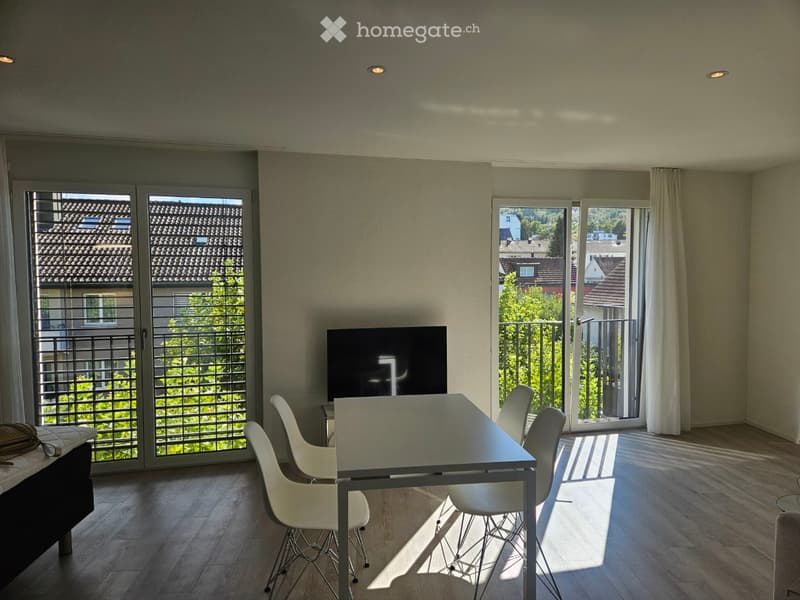 Bright dining area with a modern glass table, white chairs, and a large screen, with large windows and a view of the neighborhood.