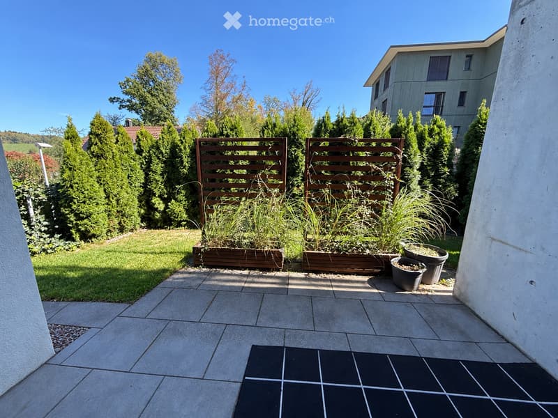 A well-maintained garden area with a paved patio, wooden planters, and surrounding greenery, including tall coniferous trees and shrubs. The area is enclosed by a wooden fence and features a modern building in the background.