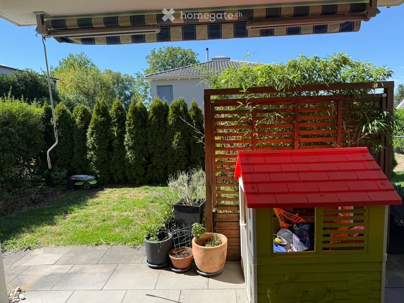 Garden area with a wooden fence, potted plants, a small toy house, and a covered patio area with an awning.