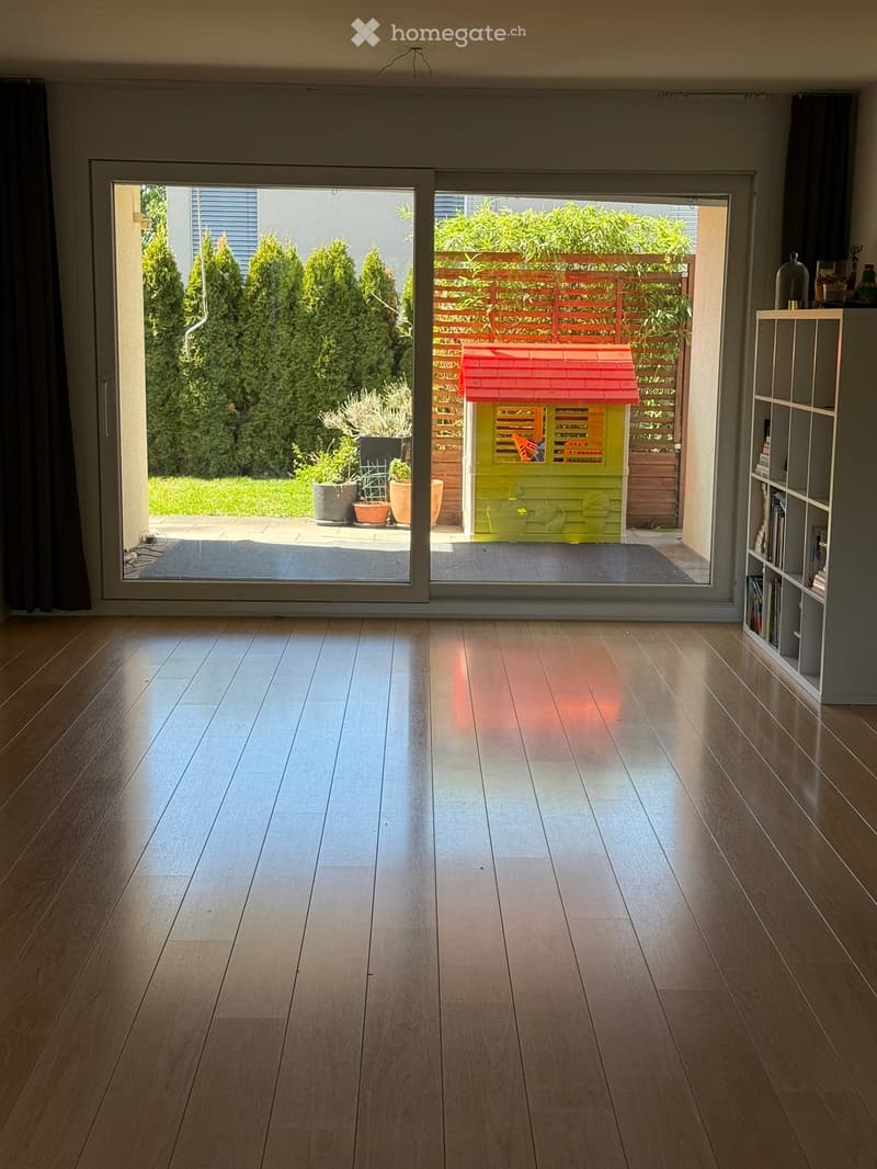 Empty room with wooden flooring, large sliding glass doors leading to a garden area with a playhouse, potted plants, and trees.