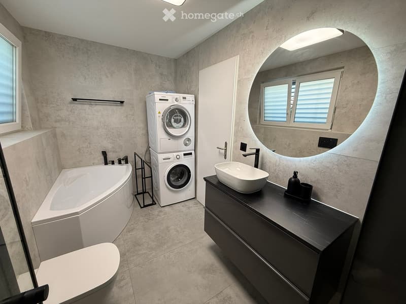 Modern bathroom with a bathtub, toilet, sink, and a stacked washing machine. Features include a large mirror, sleek black vanity, and contemporary fixtures.