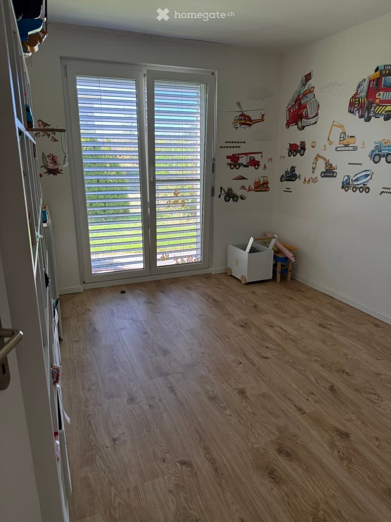 Empty room with wooden flooring, large window with blinds, and wall decals featuring vehicles and construction equipment.