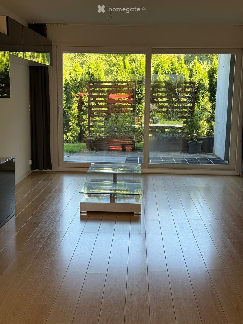 Bright, minimalist living room with wooden flooring, large sliding glass doors opening to a garden, and a sleek glass coffee table.