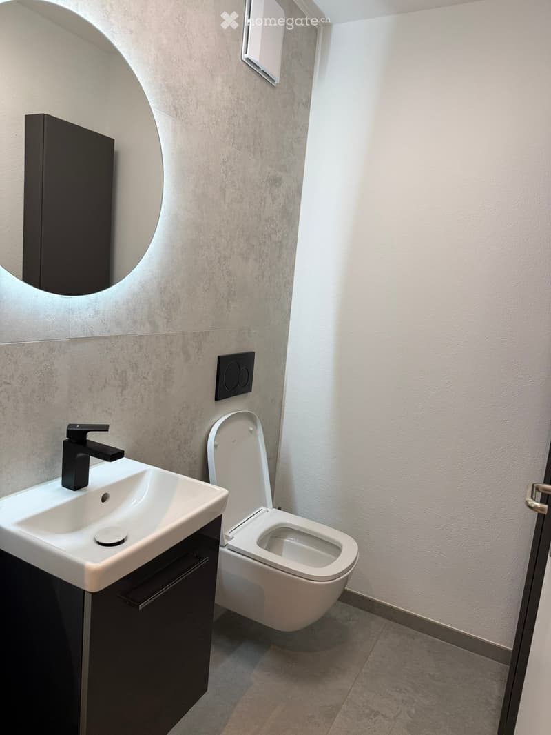 Modern bathroom with a white sink, black faucet, toilet, and a round mirror on the wall.
