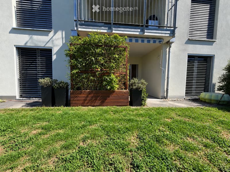 Well-maintained garden with green grass, wooden planter box, potted plants, and a small balcony with a striped awning above.