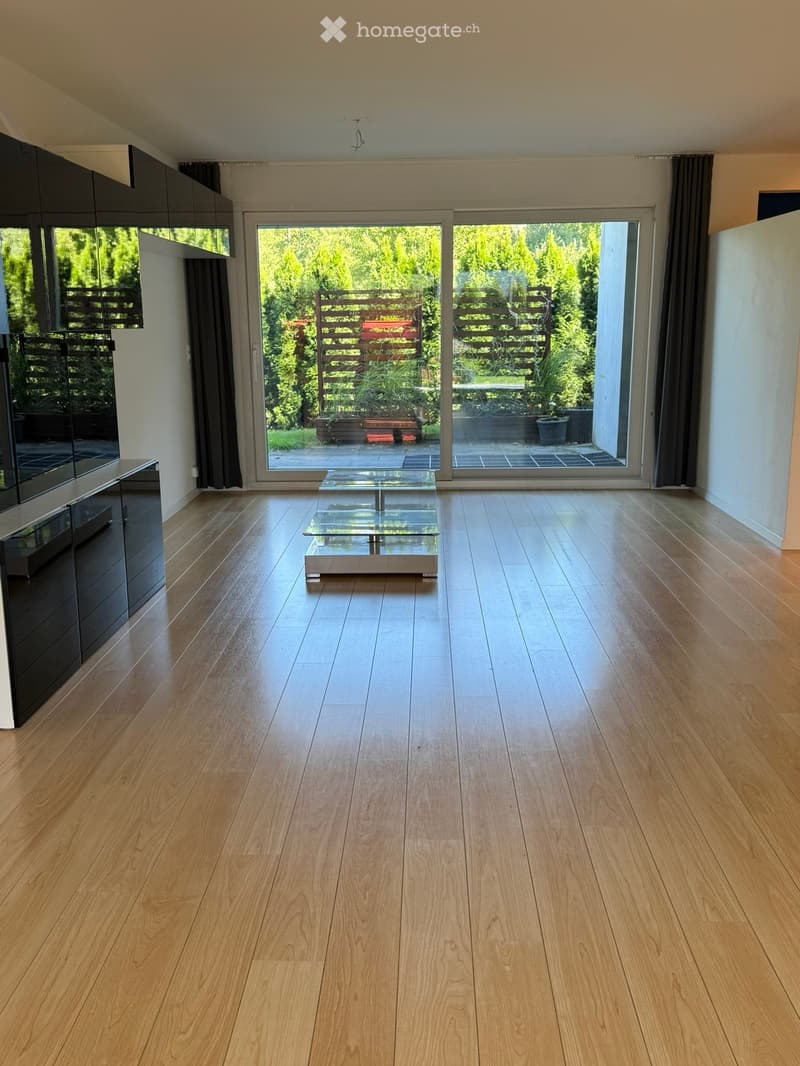 Bright, open living space with wooden flooring, large sliding glass doors leading to a garden, and a minimalist glass coffee table.