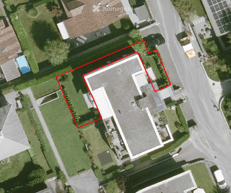Aerial view of a residential building with a flat roof, surrounded by a well-maintained lawn and trees. The building is outlined in red, indicating a property boundary.