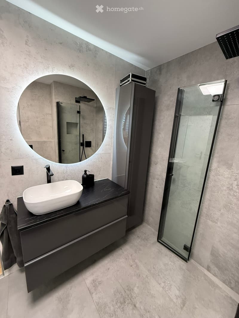 Modern bathroom with a white sink, black countertop, large round mirror, glass shower enclosure, and sleek fixtures.