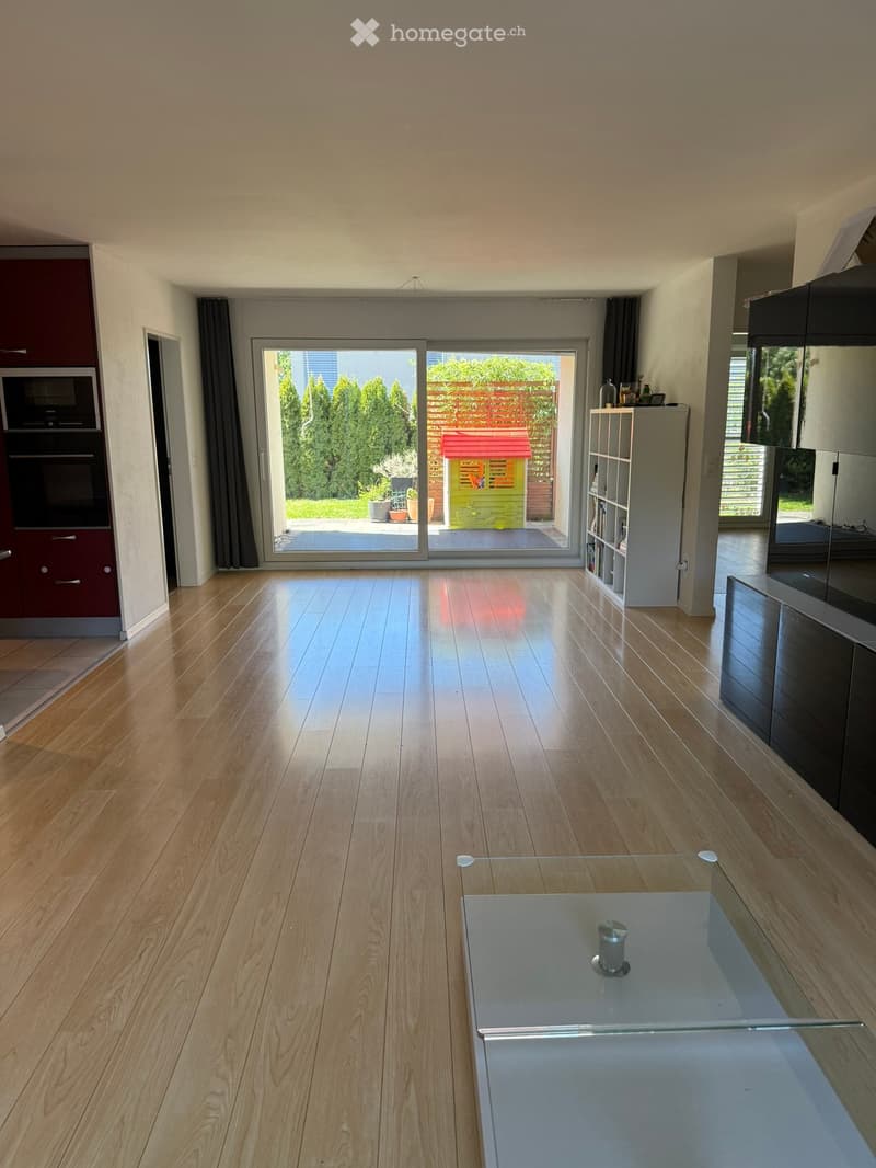 Bright, open living space with wooden flooring, large sliding glass doors leading to a garden, a glass coffee table, a white shelf, and modern kitchen appliances including a microwave and oven.