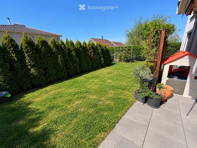 Well-maintained garden with a neatly trimmed lawn, several potted plants, a small playhouse, and a wooden trellis structure. The area is bordered by tall evergreen hedges and a paved patio.