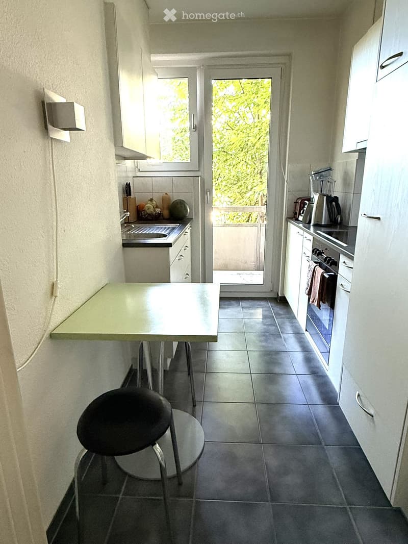 Modern kitchen with white cabinets, dark tiled floor, and a small breakfast bar with a stool. Appliances include an oven, fridge, and sink. A window and a door lead to a balcony.